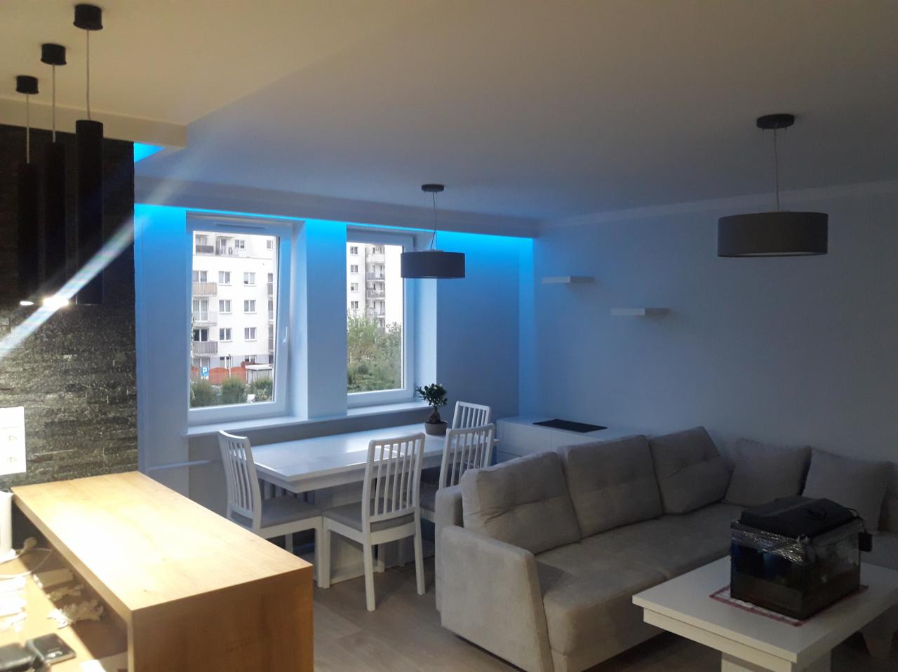 Jasne, nowocześnie urządzone wnętrze salonu z jadalnią, z oknami podświetlonymi niebieskim światłem LED, szarym narożnikiem i stołem z krzesłami.