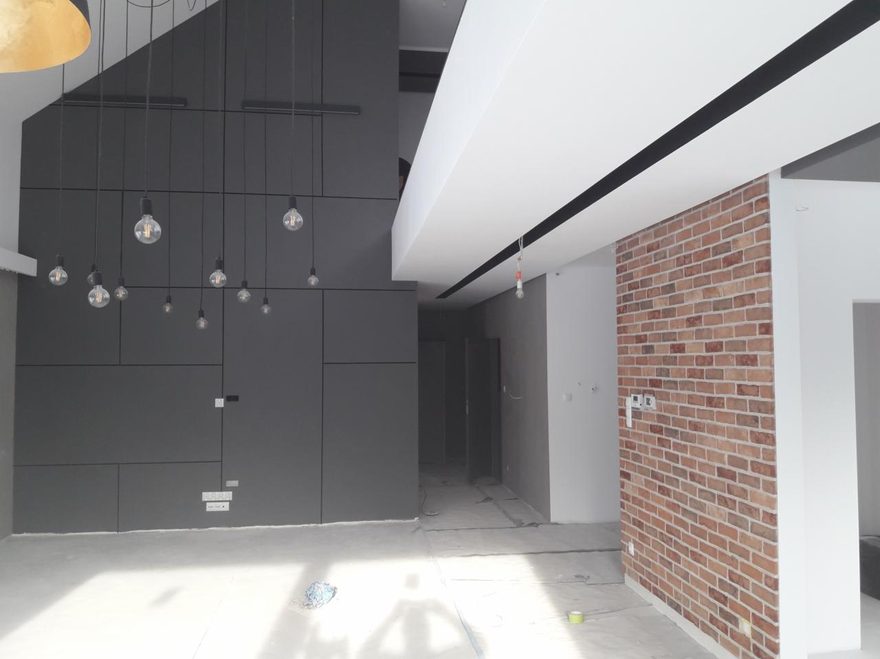 Wnętrze w trakcie wykańczania: szara ściana z geometrycznym układem paneli i wiszącymi żarówkami, fragment ściany z cegły klinkierowej, surowa podłoga zabezpieczona folią.
