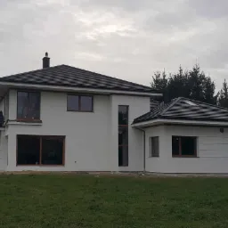 Nowoczesny, dwukondygnacyjny dom z białymi ścianami i ciemną dachówką, widoczny z perspektywy trawnika w pochmurny dzień.