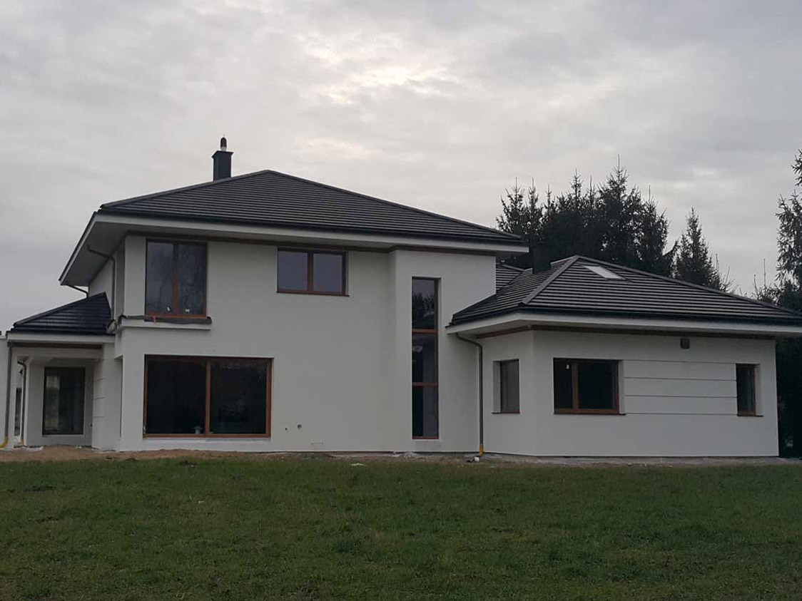 Nowoczesny, dwukondygnacyjny dom z białymi ścianami i ciemną dachówką, widoczny z perspektywy trawnika w pochmurny dzień.