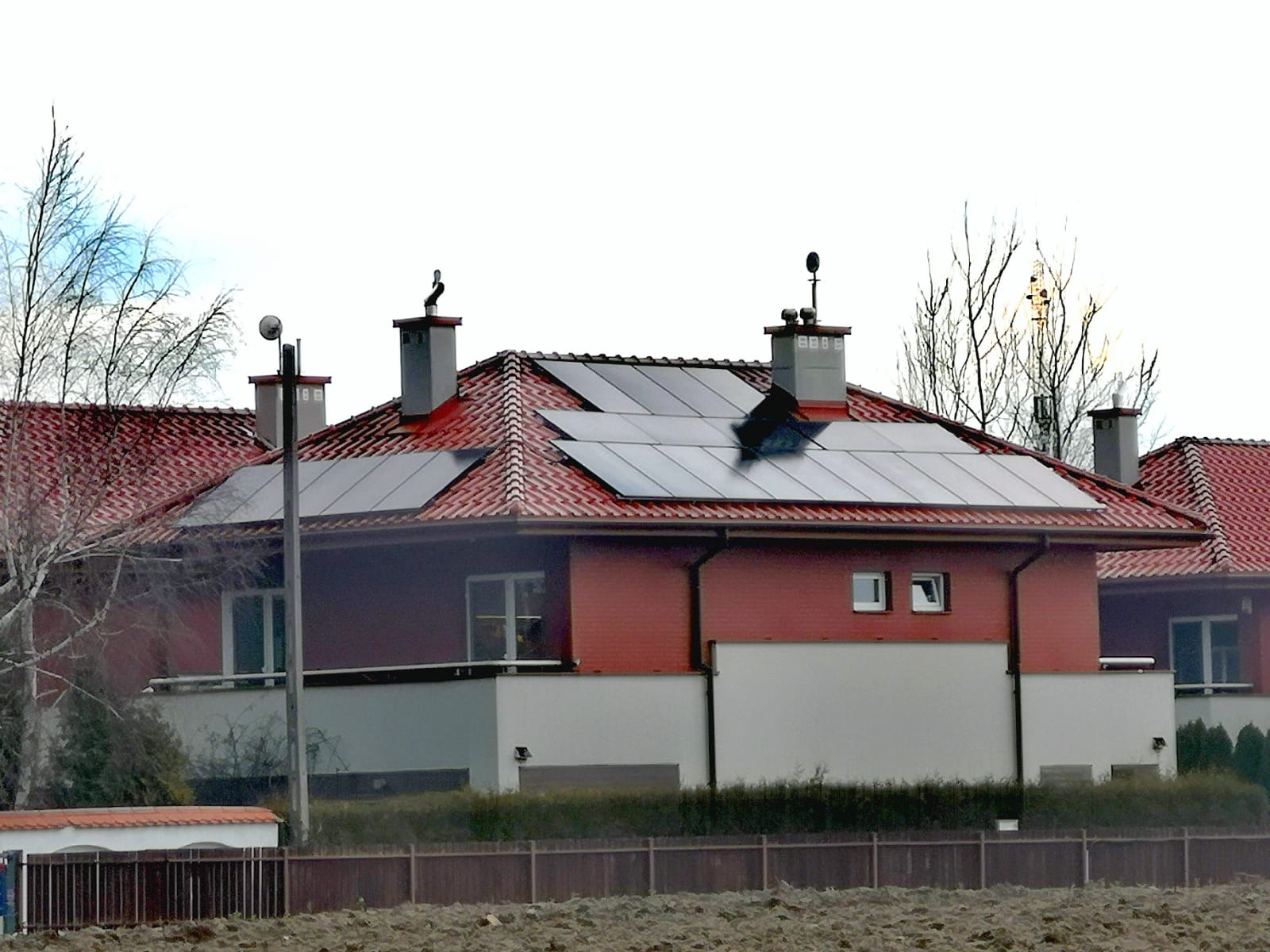 Dom jednorodzinny z czerwoną dachówką i panelami słonecznymi na dachu, widoczne kominy i antena, w tle inne budynki i drzewa bez liści.