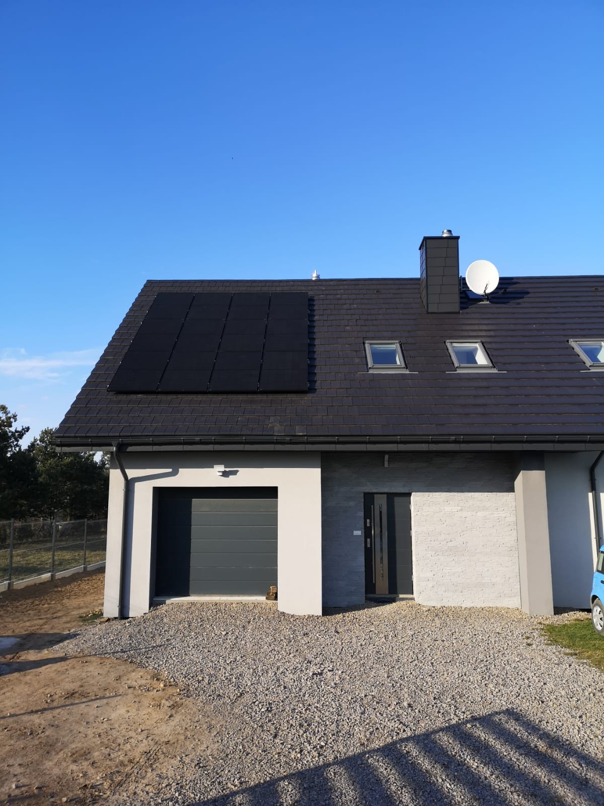 Nowoczesny dom jednorodzinny z panelami słonecznymi na dachu, widoczne okna dachowe, antena satelitarna i podjazd wysypany żwirem.