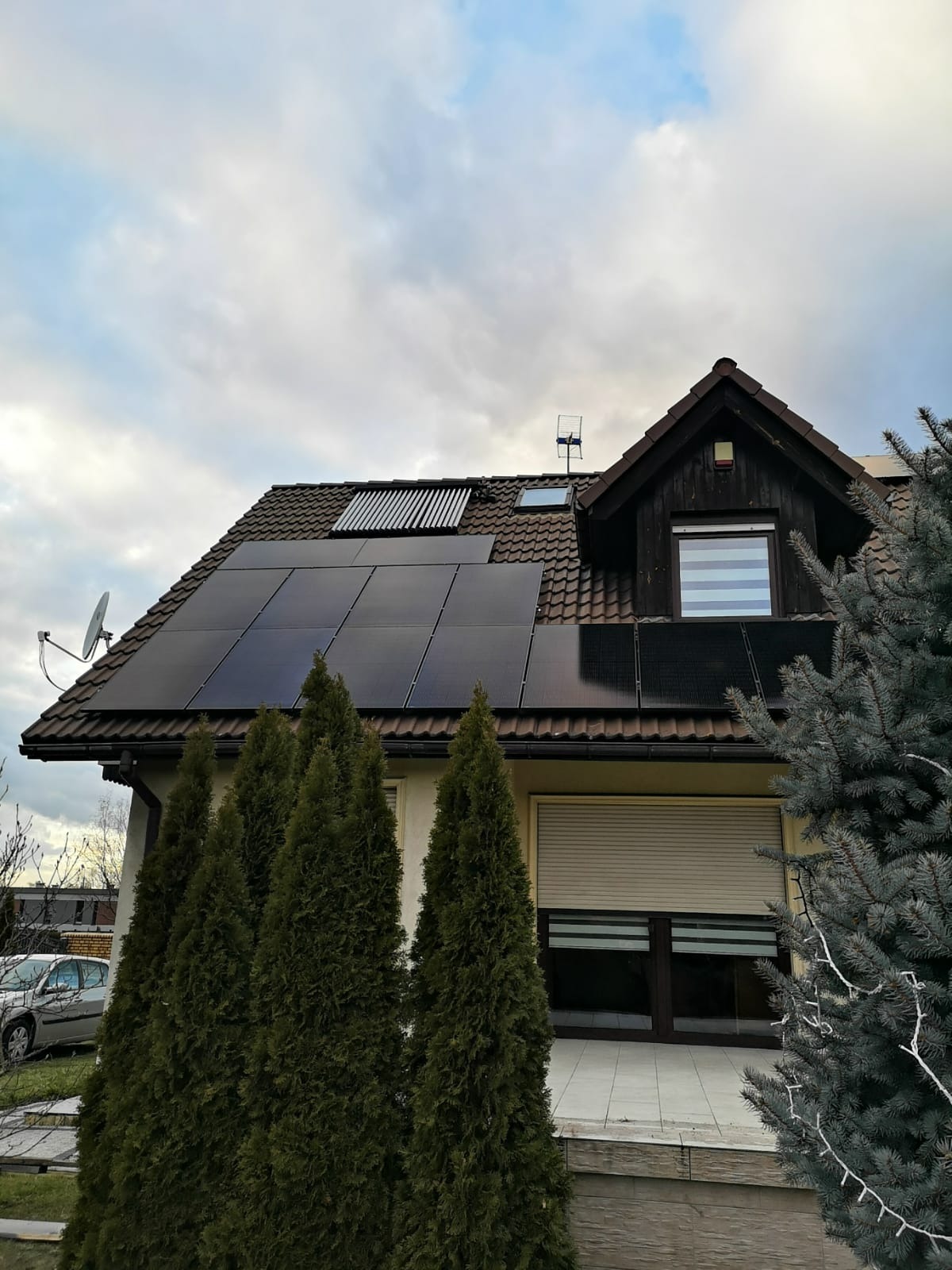 Dom jednorodzinny z panelami słonecznymi i kolektorami słonecznymi na dachu, widoczny komin, antena telewizyjna oraz rząd wysokich tuj na pierwszym planie, pochmurne niebo.