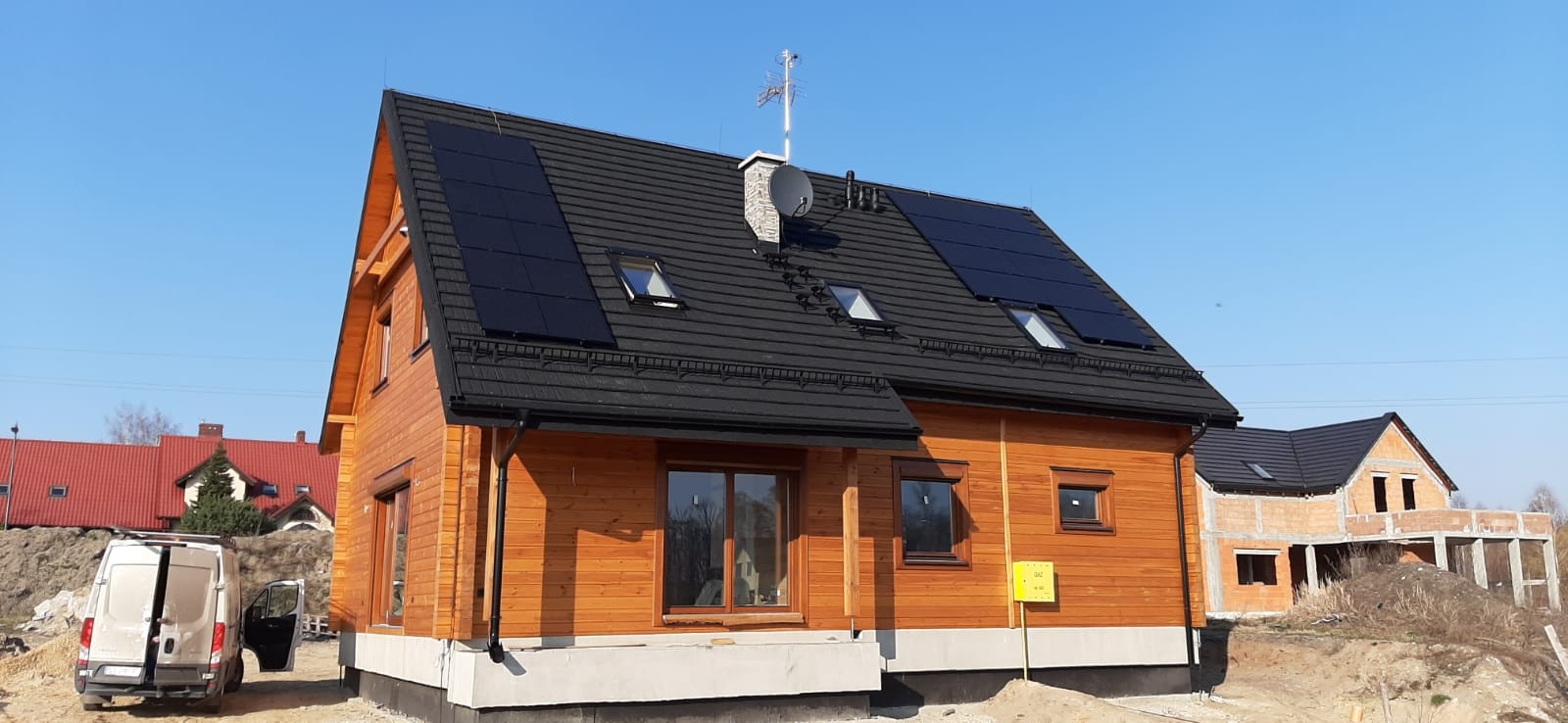 Drewniany dom z czarnym dachem i panelami słonecznymi, widoczny biały van z otwartymi drzwiami, dom w stanie surowym w tle.