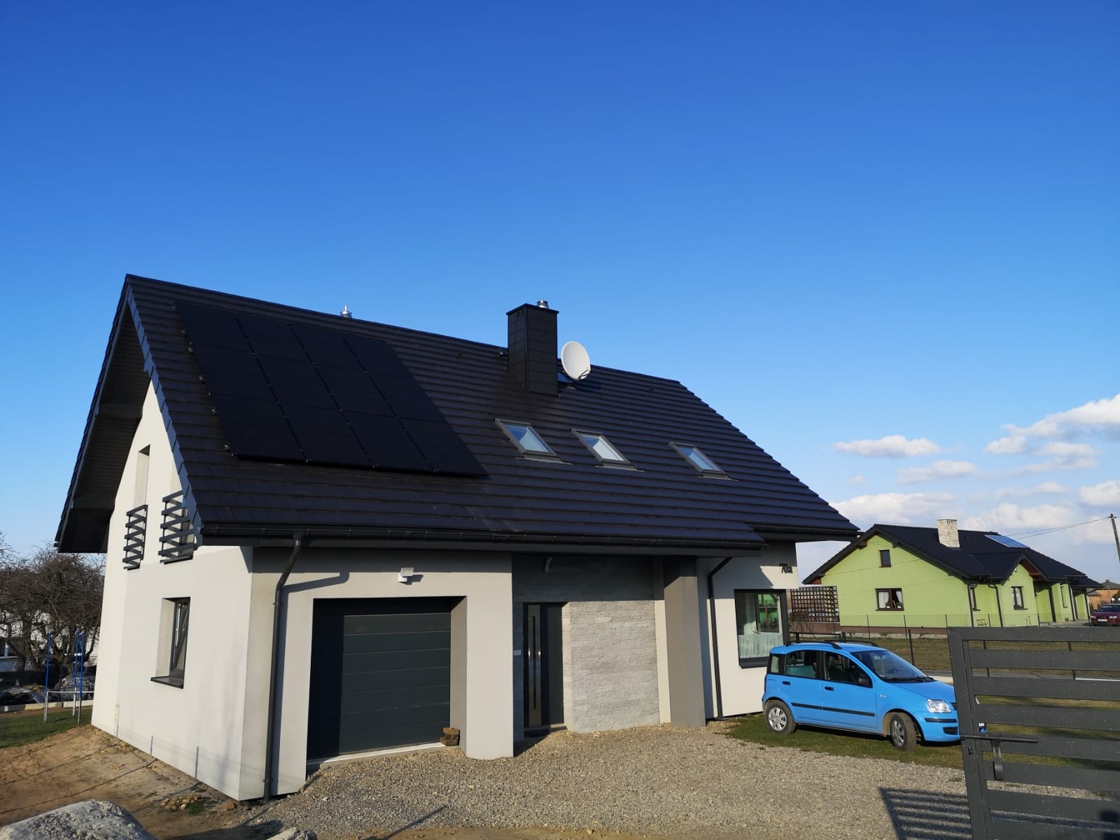 Nowoczesny dom jednorodzinny z ciemną dachówką i panelami fotowoltaicznymi na dachu, widoczne trzy okna dachowe i antena satelitarna, błękitne auto zaparkowane obok.