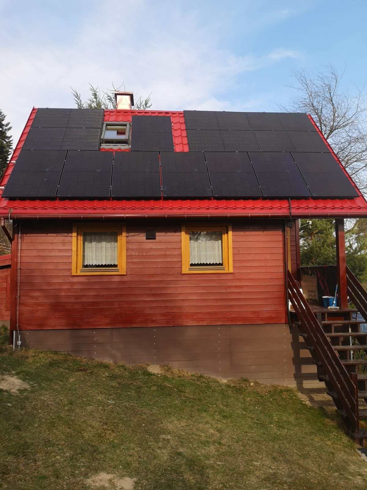 Czerwony dom jednorodzinny z zamontowanymi panelami fotowoltaicznymi na dachu, widoczne okno dachowe i schody wejściowe.