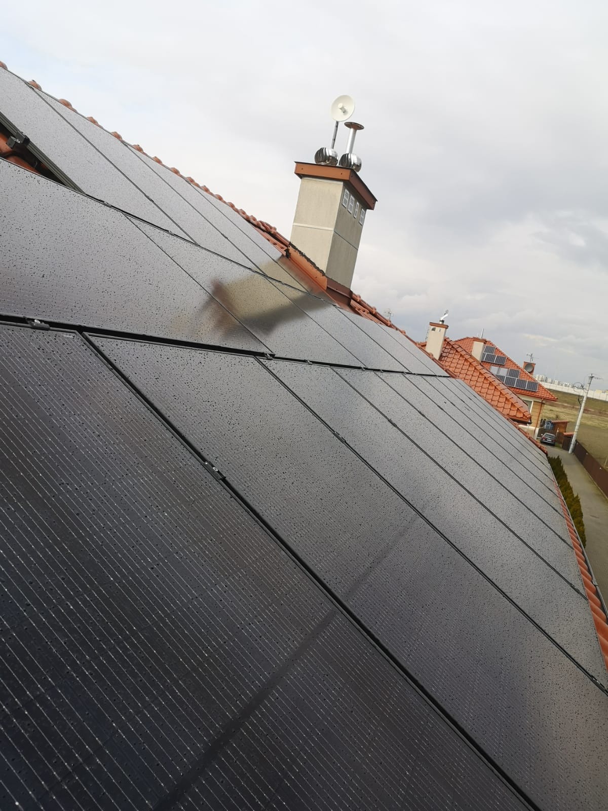 Panele słoneczne na dachu domu jednorodzinnego pokryte kroplami deszczu, widoczny komin z antenami i fragment sąsiednich budynków w tle.