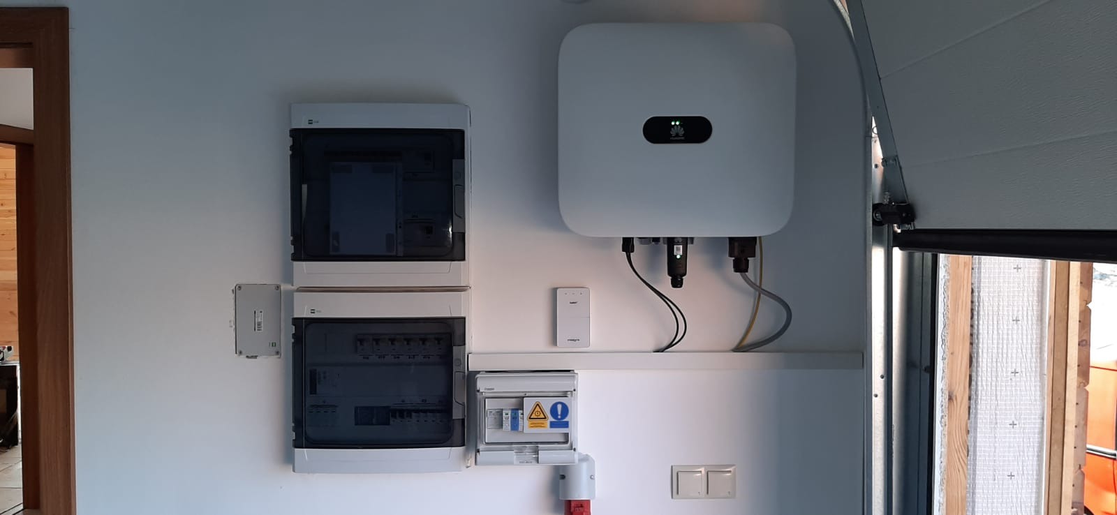 Instalacja wewnętrzna: inwerter marki Huawei, rozdzielnice elektryczne oraz moduł komunikacyjny na białej ścianie w garażu z częściowo otwartą bramą.