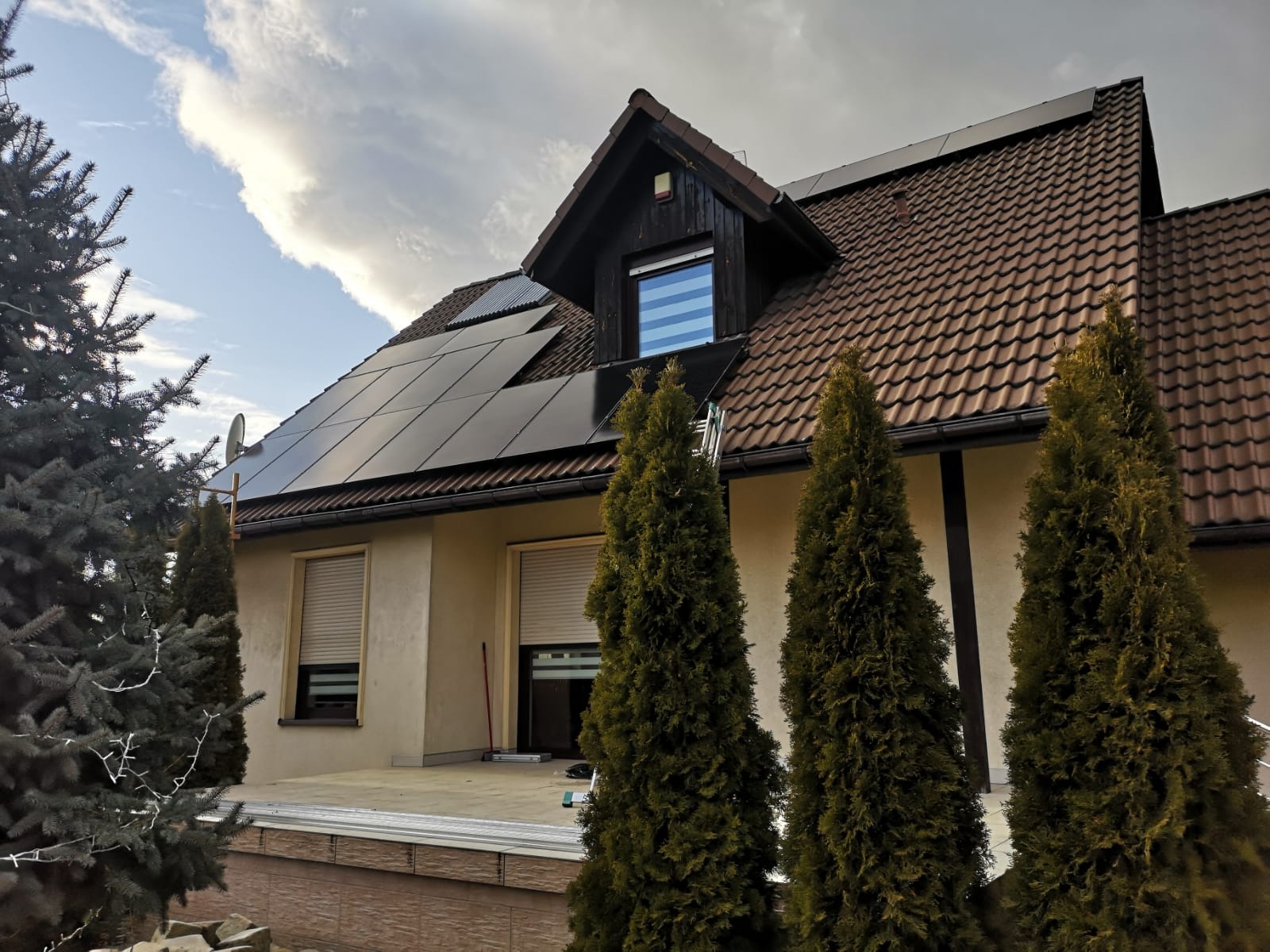 Dom jednorodzinny z zamontowanymi panelami słonecznymi na dachu, widoczne drabina i elementy montażowe na tarasie.