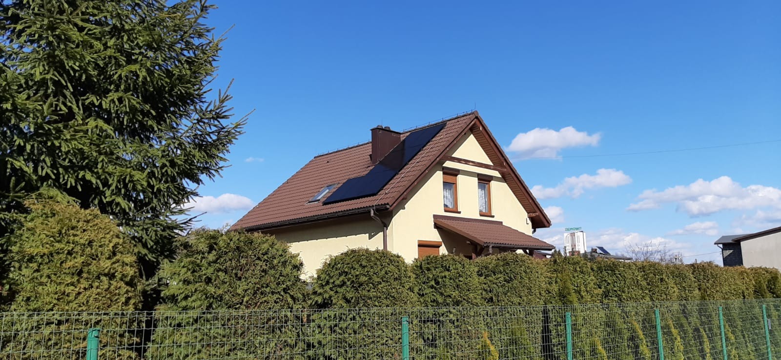 Jednorodzinny dom z brązowym dachem i panelami słonecznymi na dachu, widok zza zielonego żywopłotu i metalowej siatki ogrodzeniowej w słoneczny dzień.