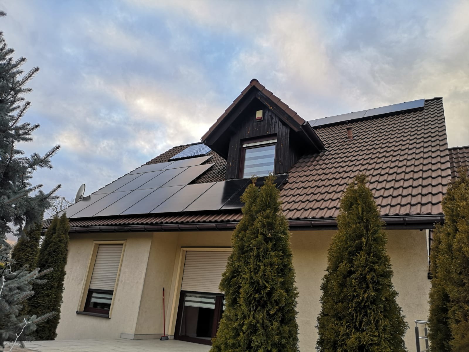 Dom jednorodzinny z brązową dachówką i panelami słonecznymi na dachu, widok z dołu w pochmurny dzień.