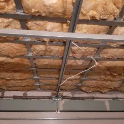 Montaż wełny mineralnej w metalowym stelażu sufitu podwieszanego, widoczna instalacja elektryczna, fragment bramy garażowej w tle.