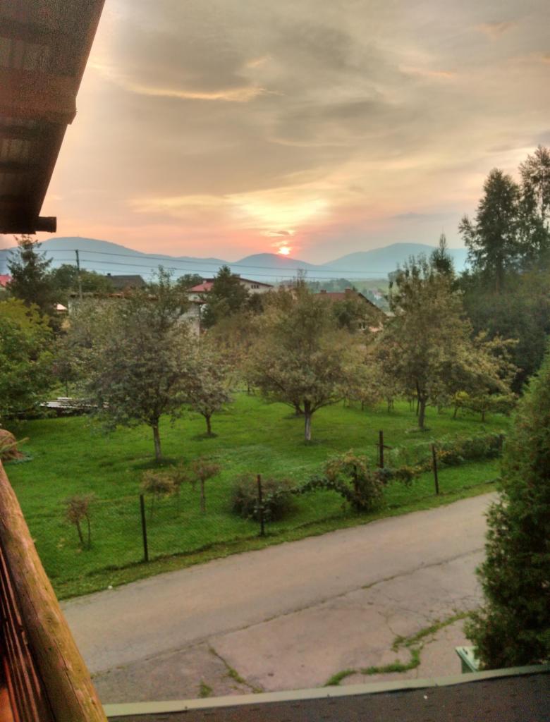 Widok z balkonu na zielony trawnik z drzewami, drogę i góry w oddali podczas zachodu słońca.