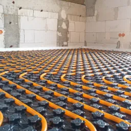 Pomarańczowe rury ogrzewania podłogowego ułożone na czarnej macie montażowej w surowym wnętrzu z widocznymi bloczkami i drewnianą konstrukcją dachu.