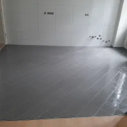 Ułożona szara, błyszcząca podłoga z płytek w pustym pomieszczeniu z białymi ścianami, widoczne przygotowane instalacje na ścianie i listwa przypodłogowa.