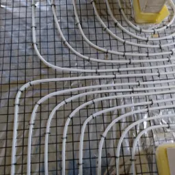 Białe rury ogrzewania podłogowego ułożone na czarnej siatce z folią aluminiową w tle, widoczne fragmenty schodów i żółtej pianki.