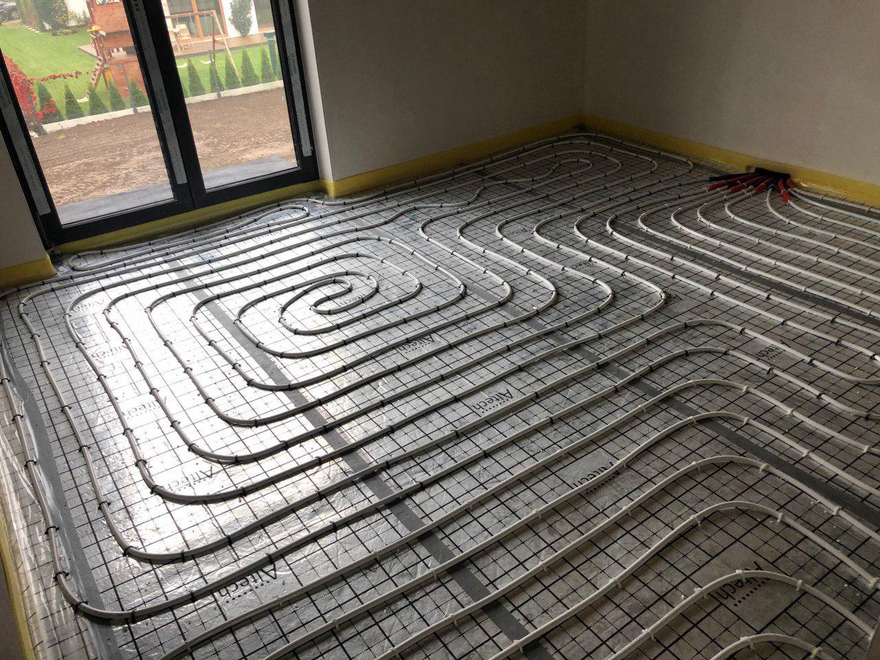 Układanie rur ogrzewania podłogowego na folii z siatką, widoczne narzędzia i taśma zabezpieczająca przy ścianach, okno z widokiem na trawnik w tle.