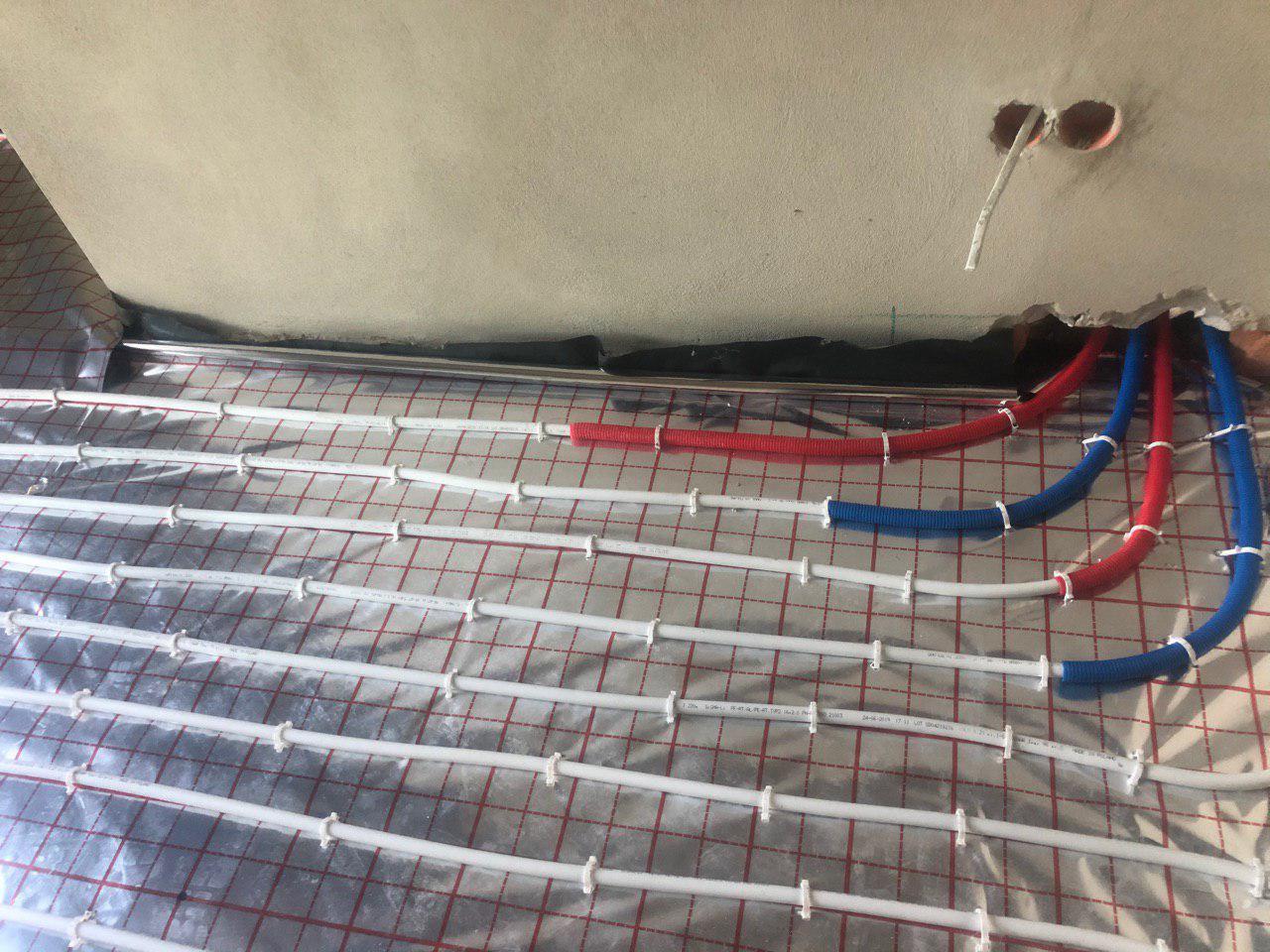 Montaż ogrzewania podłogowego: rury grzewcze ułożone na folii izolacyjnej z widocznymi rurami doprowadzającymi w kolorze czerwonym i niebieskim, surowy stan wykończenia.