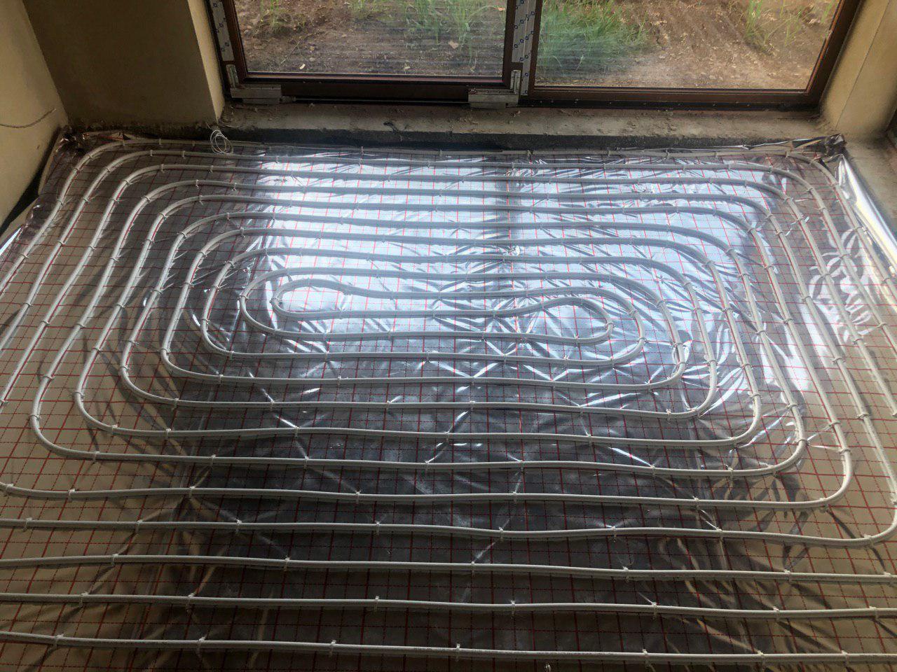 Rozłożona instalacja ogrzewania podłogowego na folii izolacyjnej z siatką, widok z góry, okno w tle.