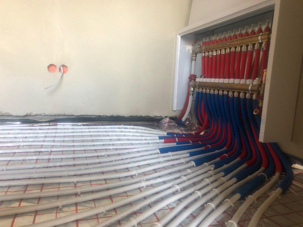 Rozłożone rury ogrzewania podłogowego na siatce, z widoczną rozdzielnicą z czerwonymi i niebieskimi rurami doprowadzającymi, surowy stan ścian z wystającymi kablami.