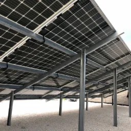 Widok od spodu na zainstalowane panele fotowoltaiczne na metalowej konstrukcji wsporczej, z widocznymi kablami i białym żwirem pod spodem.