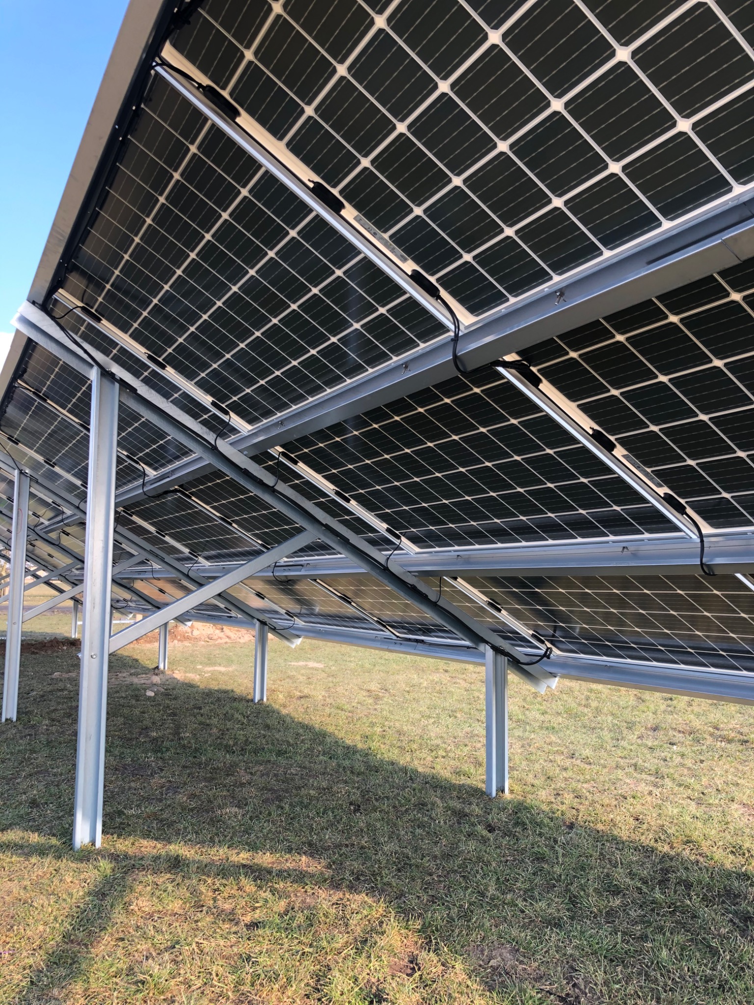 Widok od spodu na panele fotowoltaiczne zamontowane na metalowej konstrukcji, z widocznymi kablami i trawą w tle.