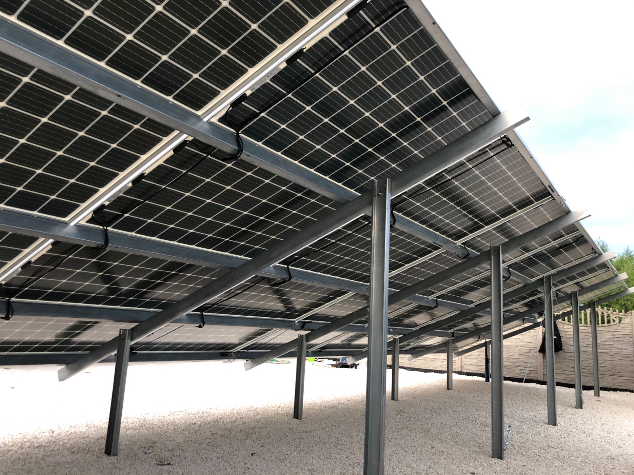 Widok od spodu na zainstalowane panele fotowoltaiczne na metalowej konstrukcji wsporczej, z widocznymi kablami i białym żwirem pod spodem.