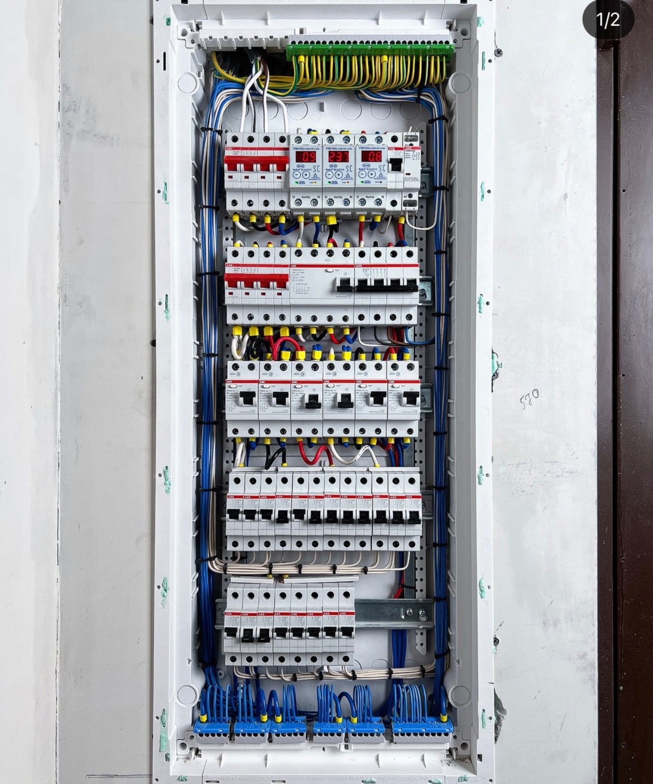 Rozdzielnica elektryczna z zamontowanymi bezpiecznikami, przewodami w kolorach niebieskim, żółtym i zielonym oraz wyświetlaczami LED, umieszczona w białej obudowie na ścianie z widocznymi śladami...