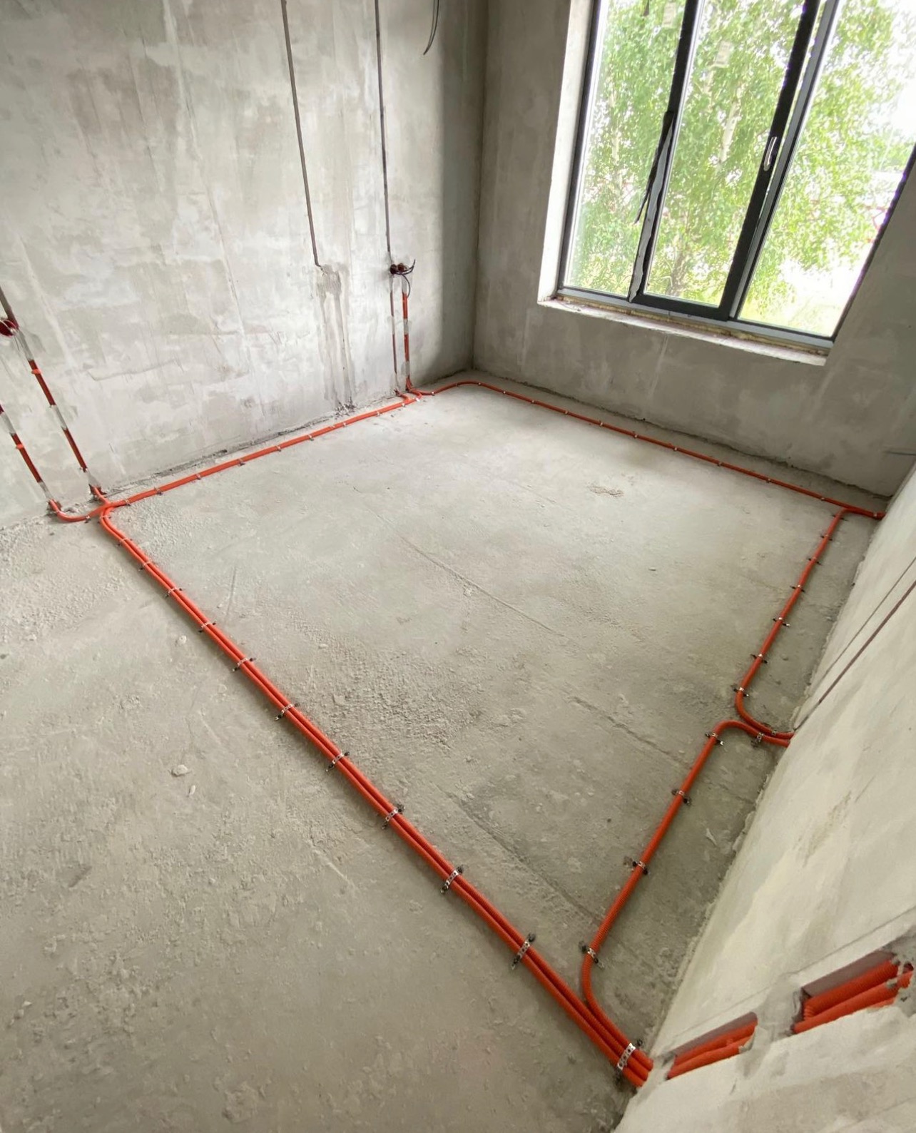 Pomieszczenie w stanie surowym z widoczną, pomarańczową instalacją elektryczną ułożoną wzdłuż ścian i pod sufitem, zabezpieczoną metalowymi uchwytami.