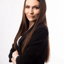 Portret kobiety w czarnym żakiecie na białym tle, skrzyżowane ręce, długie brązowe włosy, profesjonalny wizerunek.