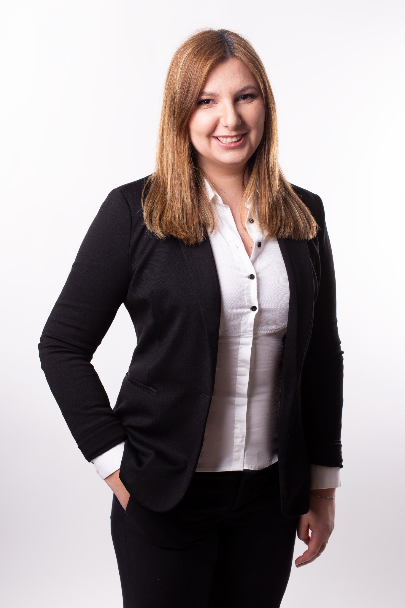 Portret uśmiechniętej kobiety w czarnym żakiecie i białej koszuli, z ręką w kieszeni, na jasnym tle.