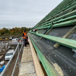 Montaż łat dachowych na membranie, widok z rusztowania, w tle plac budowy i jesienny krajobraz.