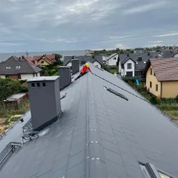 Dekarz w kamizelce odblaskowej i kasku montuje dachówkę na skośnym dachu domu jednorodzinnego z oknami dachowymi i trzema kominami, w tle zabudowa osiedlowa pod pochmurnym niebem.