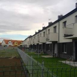 Szereg nowych domów z balkonami, świeżo założone trawniki, ogrodzenie panelowe, pochmurne niebo.