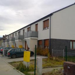 Słowiński Usługi Budowlane Michał Słowiński - Szeregówka z białą elewacją i ciemną cegłą na dole, balkony z ażurowymi barierkami, brązowe rolety w oknach, pochmurne niebo.