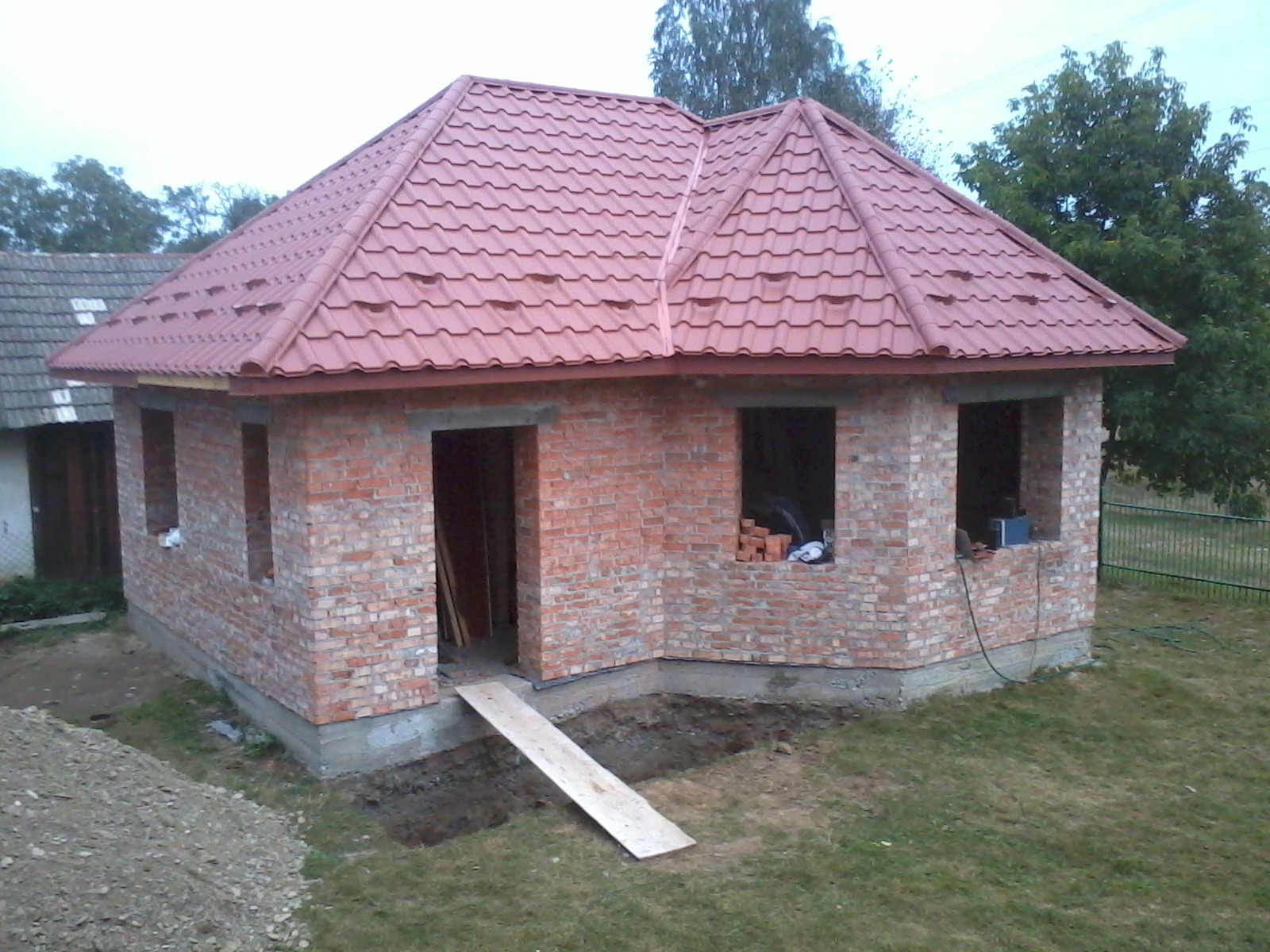 Niedokończony budynek z czerwonej cegły z nowym dachem z blachodachówki w kolorze ceglastym; widoczna deska prowadząca do wejścia i stos cegieł w oknie.