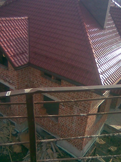 Wysoki kąt widzenia na budynek w trakcie budowy z nowym, czerwonym dachem z dachówki ceramicznej, widoczna surowa cegła i elementy konstrukcyjne.