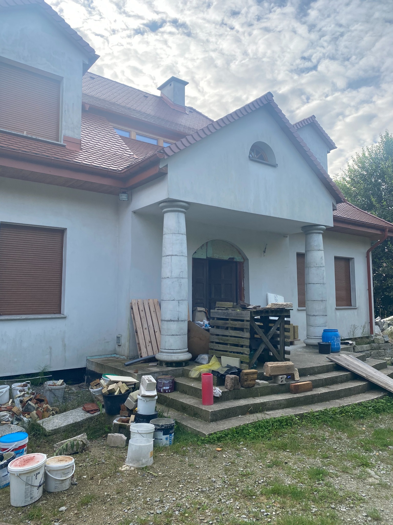 Dom jednorodzinny w trakcie prac elewacyjnych, widoczne rusztowania, materiały budowlane i niedokończona elewacja w kolorze białym, z elementami dekoracyjnymi w postaci kolumn.