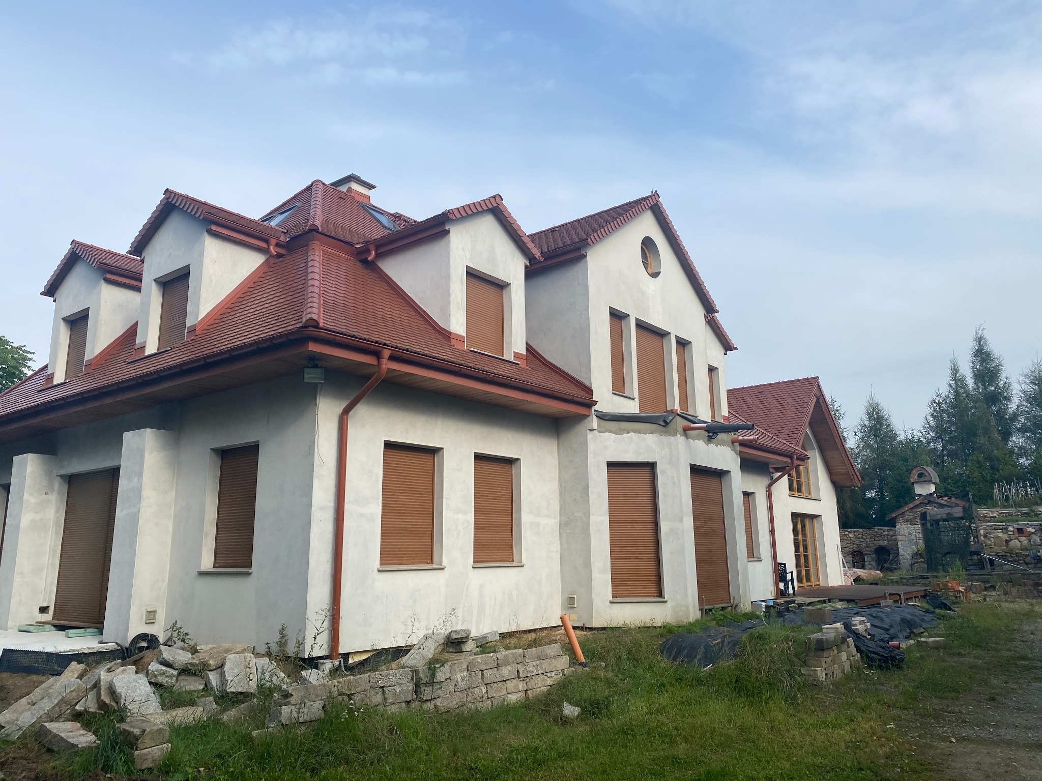 Niedokończony dom z szarą elewacją i czerwoną dachówką, widoczne rusztowania i materiały budowlane na trawniku.