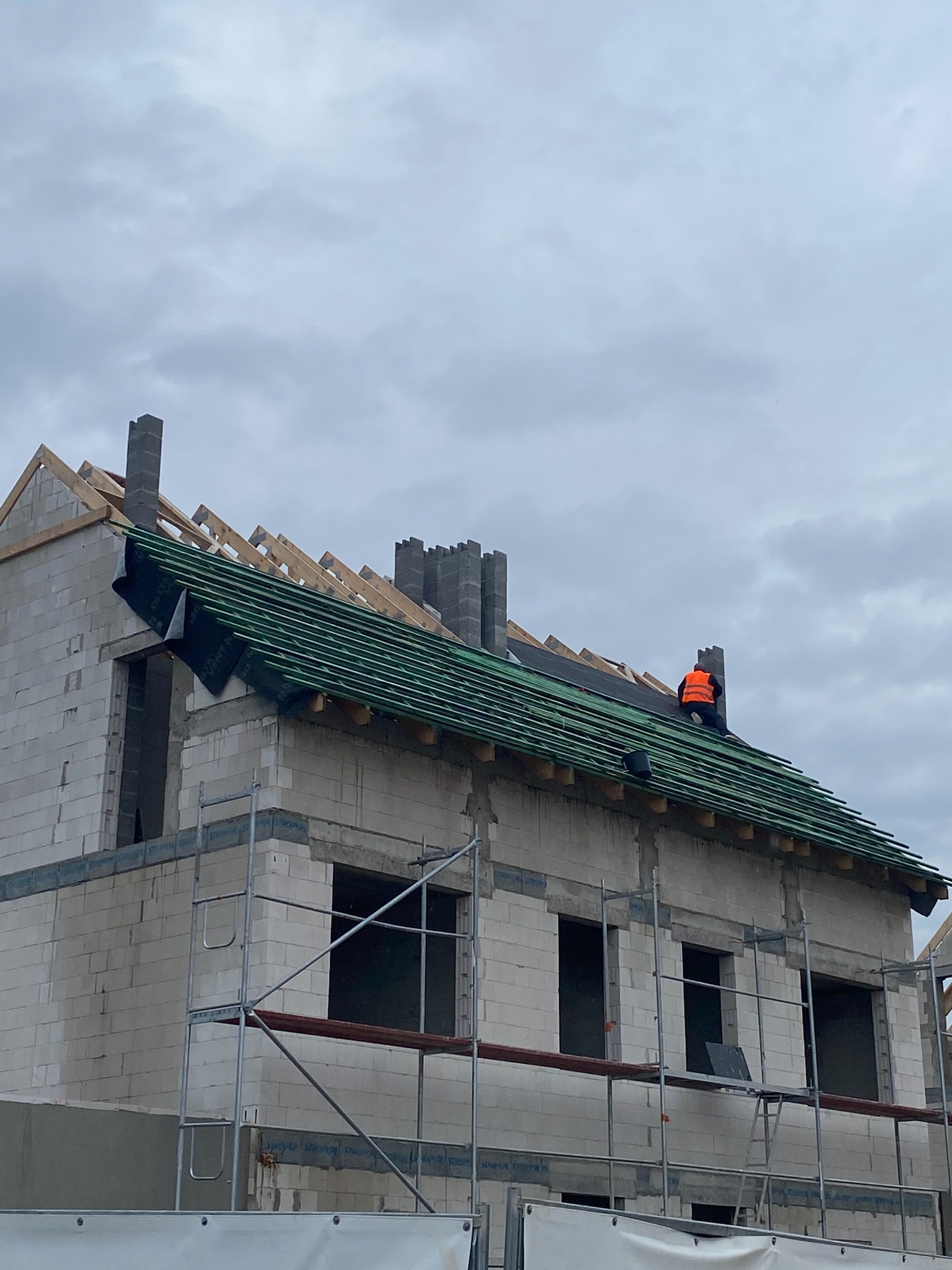 Budowa dachu domu jednorodzinnego: widok na konstrukcję więźby dachowej z deskowaniem i membraną, pracownik w pomarańczowej kamizelce na dachu, rusztowania wokół budynku.