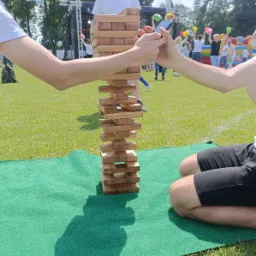 Gra w Jengę na trawie podczas imprezy plenerowej. W tle widoczna scena i bawiący się ludzie. Uczestnicy delikatnie wyciągają drewniane klocki z wieży.