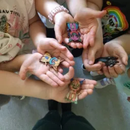 Dzieci prezentują ręcznie robione, kolorowe przypinki z motywami fantastycznych stworzeń, trzymając je na otwartych dłoniach, widok z góry.