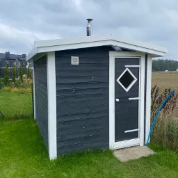 Mała, ciemna budowla z drewnianych desek z białymi wykończeniami, prawdopodobnie sauna lub domek ogrodowy, z kominem na dachu i małym okienkiem w kształcie rombu w drzwiach, stoi na trawniku.