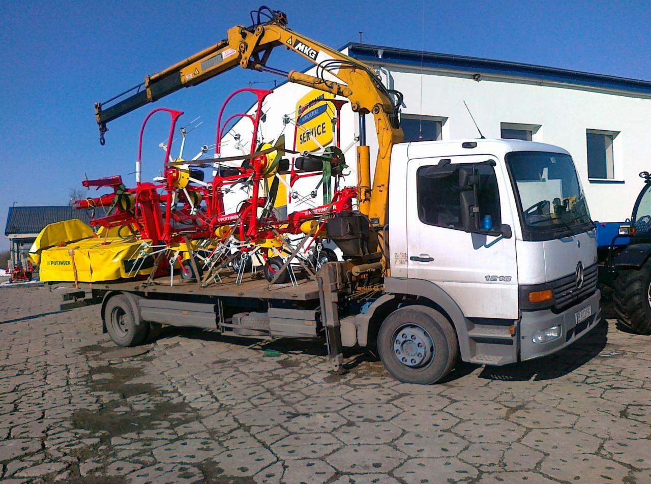 Ciężarówka z dźwigiem przewozi maszyny rolnicze marki Pöttinger. Widoczny budynek z napisem 'Agriculture Service'.