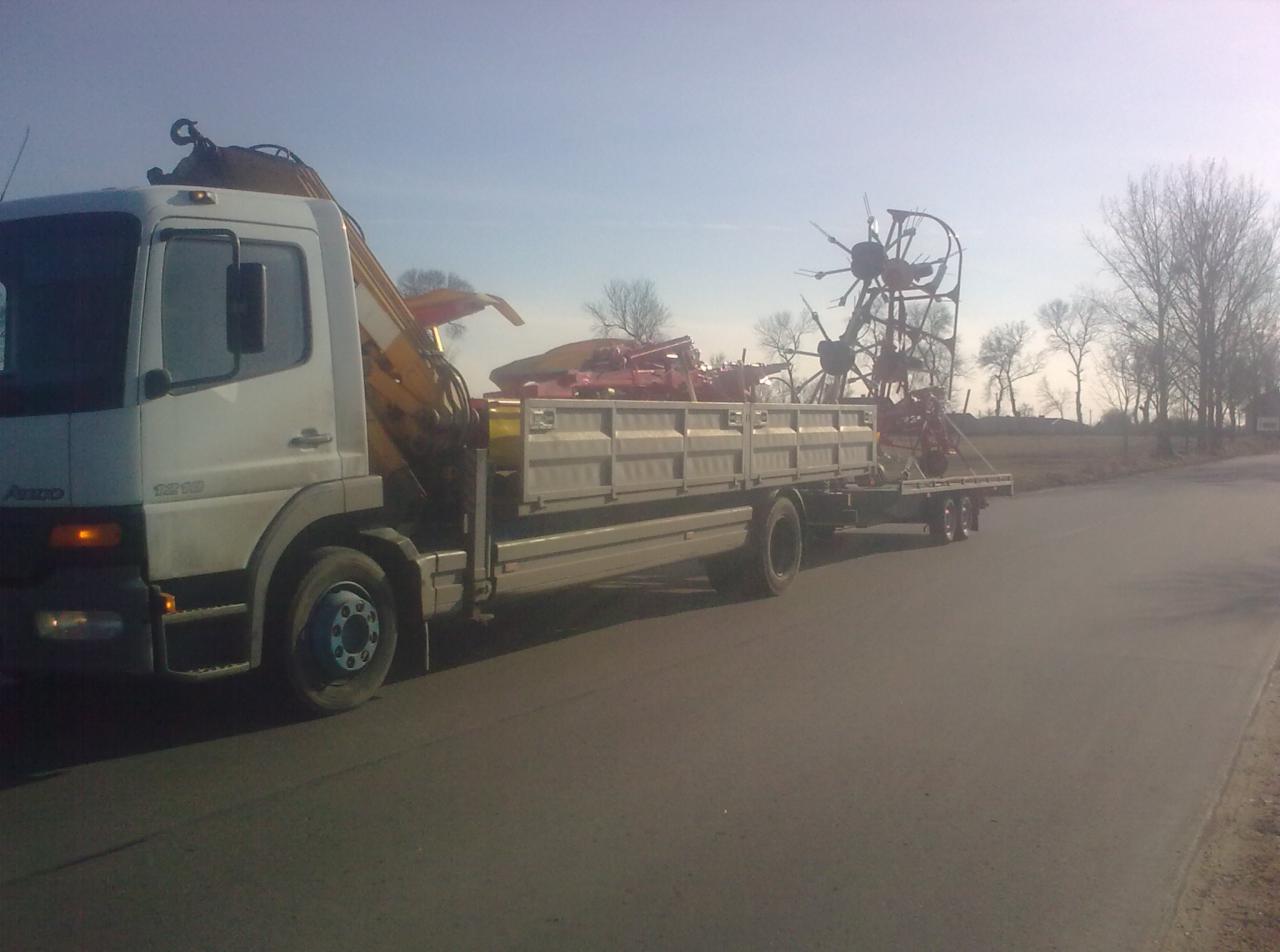 Ciężarówka z żółtym dźwigiem przewozi maszyny rolnicze na przyczepie. Transport po asfalcie w słoneczny dzień.
