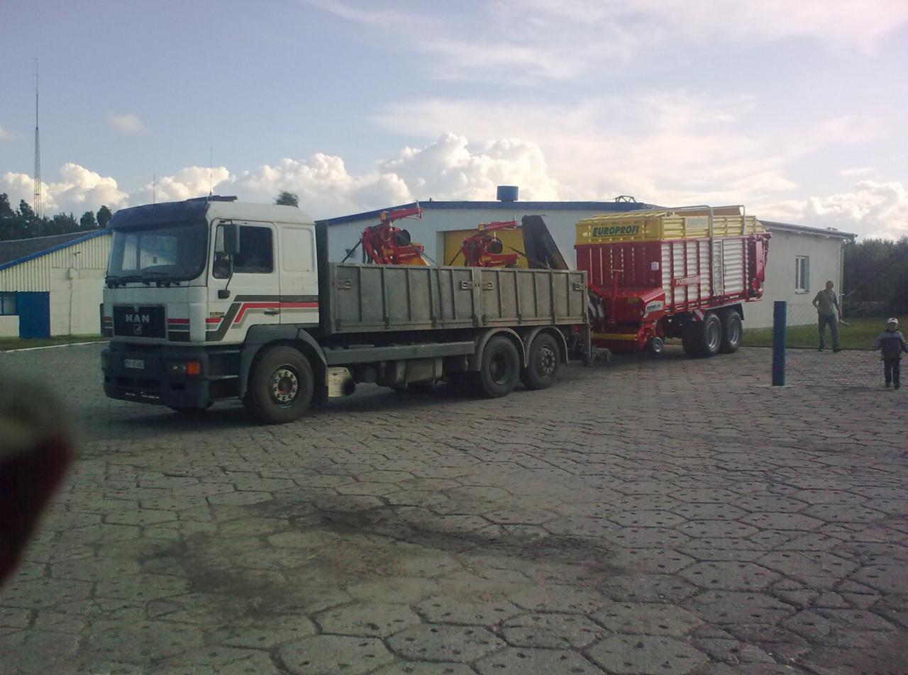 Ciężarówka MAN z przyczepą rolniczą Europrofi na brukowanym placu, w tle budynki i osoby. Transport maszyn rolniczych.