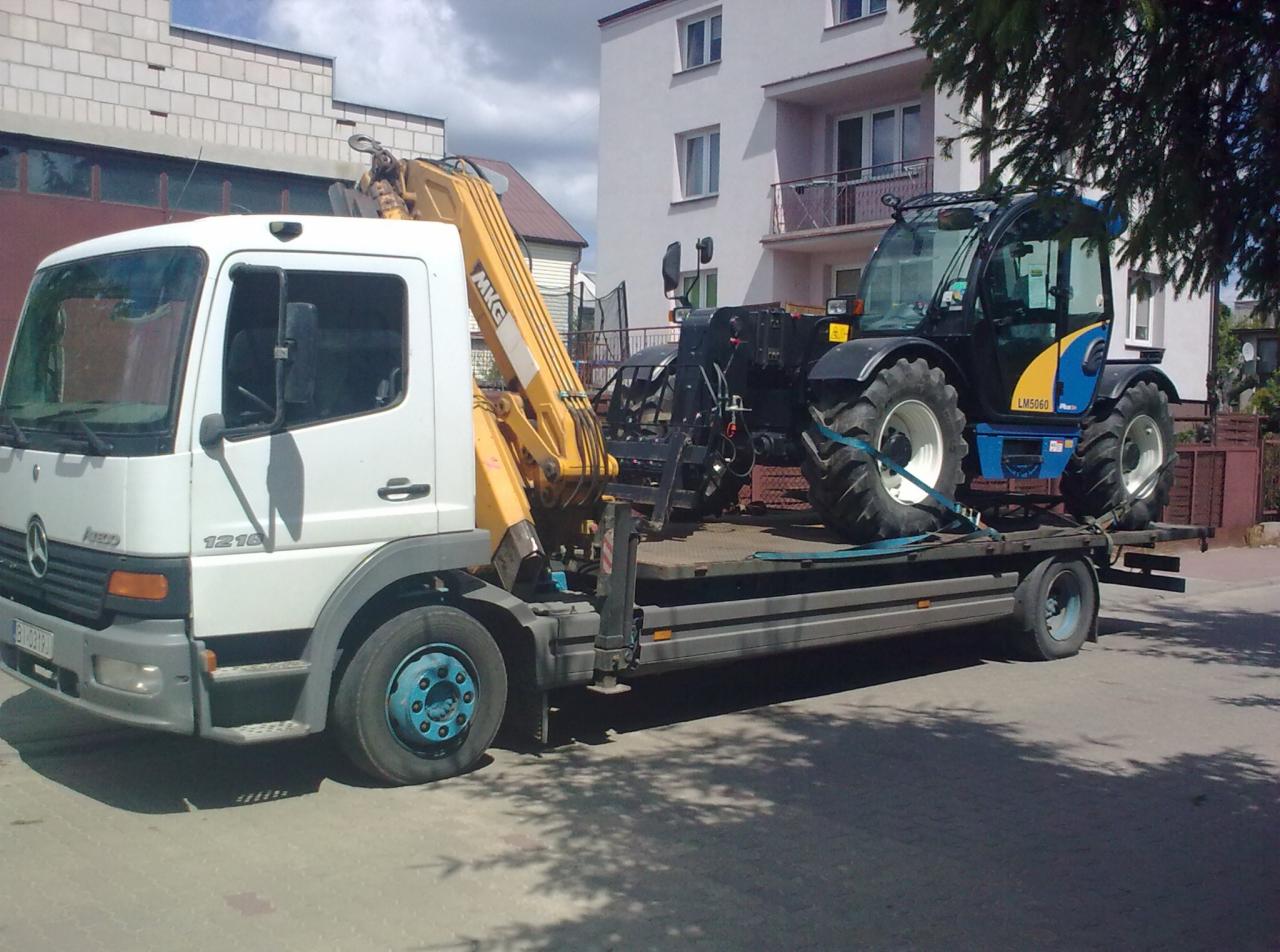Ciężarówka z żółtym dźwigiem przewozi niebiesko-czarny traktor rolniczy na niskopodwoziowej platformie. Budynek w tle.