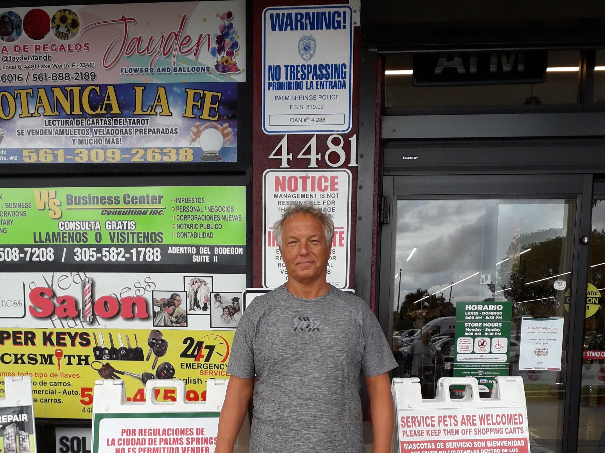 Mężczyzna na tle witryny sklepowej w Łodzi, z szyldami i ogłoszeniami w języku angielskim i hiszpańskim. Widoczne napisy 'Keys', 'Business Center', 'Warning!'.