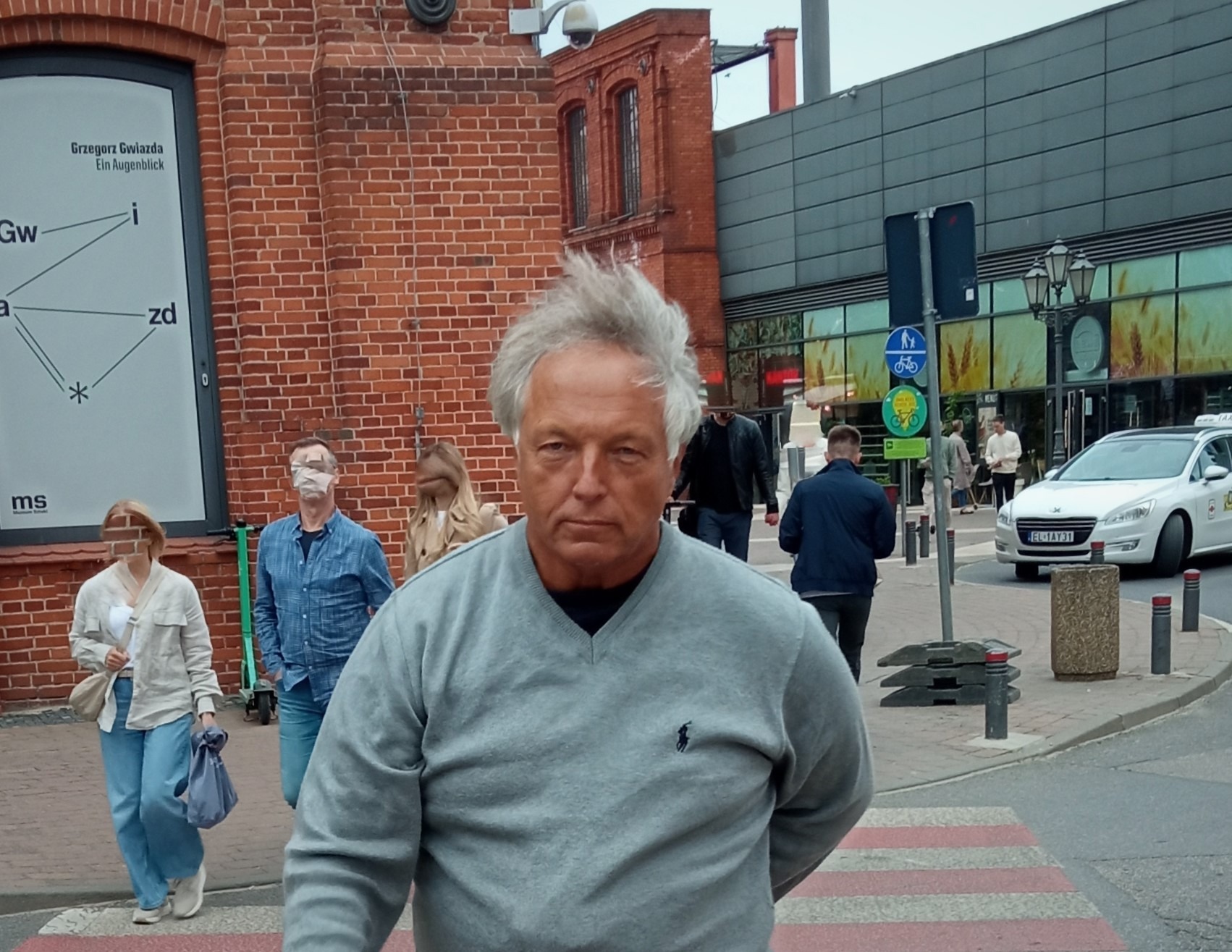 Mężczyzna w szarym swetrze na tle ceglanej architektury i przechodniów na pasach. Widoczne znaki drogowe i taksówka. Ujęcie z dołu, lekko zniekształcona perspektywa.