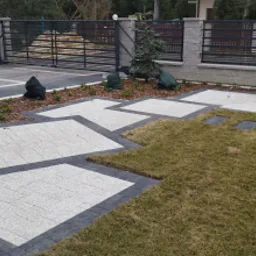 Ułożona jasnoszara kostka brukowa z ciemnoszarą obwódką tworząca geometryczny wzór na trawniku, w tle brama wjazdowa i ogrodzenie z szarego kamienia łupanego.